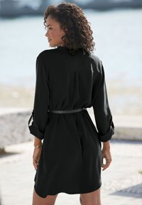Robe noire à ceinture, avec des manches trois quarts, une coupe décontractée et un dos plissé, mise en valeur sur un fond extérieur.