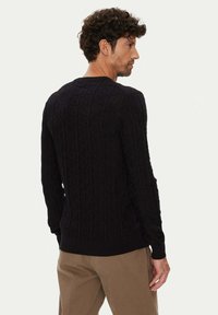 Schwarzer gestrickter Pullover mit strukturiertem Muster, Rundhalsausschnitt und geripptem Saum, getragen über beigen Hosen. Rückansicht sichtbar.