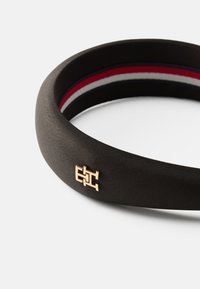 Fekete anyagú fejpánt sima textúrával, arany fém logóval és belső piros, fehér és tengerészkék csíkokkal.