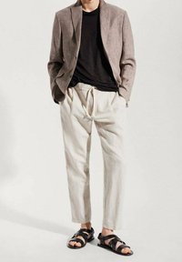 Outfit da uomo composto da un blazer in lino beige, t-shirt nera, pantaloni crema con coulisse e sandali neri con fasce. Design semplice e colori chiari.