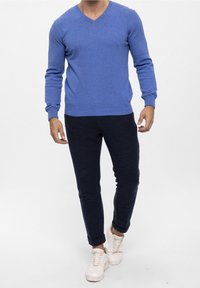 Homme portant un pull bleu à col en V, un pantalon texturé foncé et des baskets blanches, debout devant un fond blanc uni.