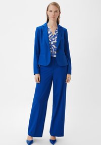 Blauer taillierter Blazer und passende weit geschnittene Hose, kombiniert mit einer gemusterten Bluse. Glattes Material mit Satin-Finish und scharfen Linien.