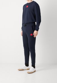 Marineblauwe sweatshirt en joggers van zachte stof, met rode merklappen. Witte sneakers met een rubberen zool maken de outfit compleet.