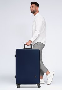 Valigia rigida blu navy con una superficie liscia, quattro ruote e manico estensibile, sostenuta da una persona che indossa pantaloni grigi e scarpe bianche.