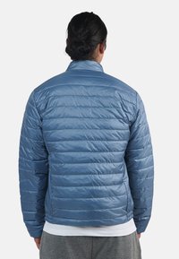 Blå puffjacka med horisontell quiltad design, hög krage och elastiska ärmslut. Slitstarkt, blanka yttertyg; lätt konstruktion.