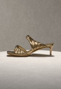 Χρυσά μεταλλικά σανδάλια με τακούνι, με υφή, πλισέ σχέδιο και ανοιχτό δάχτυλο. Διαθέτουν χαμηλό, λεπτό τακούνι και φαρδιά λωρίδα που διασχίζει το πόδι.
