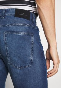 Jeans in denim blu presentano una vestibilità dritta, con una grande tasca sul retro e una patch nera con il marchio sulla vita. La texture appare leggermente sbiadita.