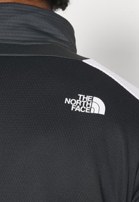 Chaqueta deportiva negra con un acabado texturizado, que presenta un cuello alto y un logo blanco en el hombro izquierdo. Detalle de franjas blancas en contraste.