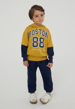 Felpa con cappuccio gialla e blu navy con testo "BOSTON 88", abbinata a pantaloni della tuta blu navy. Le scarpe sono chiare con strap in Velcro. Design semplice.