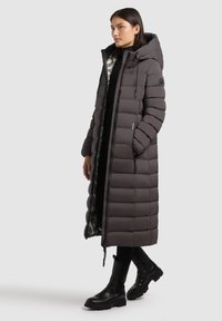 Cappotto lungo da puffa grigio scuro con cappuccio. Presenta un design trapuntato, chiusura a zip e un rivestimento argentato lucido visibile dall'apertura anteriore. Stivali neri.