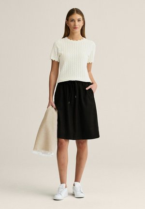 Kvinde iført en hvid kortærmet striktop, sort knælængde nederdel, hvide sneakers, holder et beige frynset tørklæde i den ene hånd.