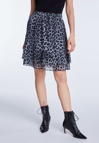 Femme portant une jupe à volants longueur genou imprimé léopard, un haut noir ajusté et des bottines à talons noires à lacets, debout devant un fond uni.