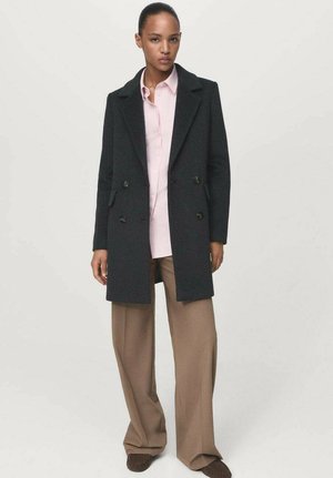 Manteau en laine noir croisé avec de larges revers, poches latérales et boutons dorés, superposé sur une chemise rose à boutons et un pantalon marron à jambes larges.