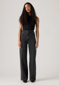 Jeans noirs taille haute, à jambes larges, en denim doux avec des poches avant et des détails de couture subtils. Portés avec un haut noir ajusté.