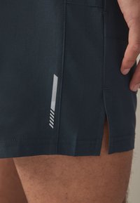 Pantaloncini sportivi blu navy con texture traspirante, spacchi laterali e dettagli riflettenti con motivo a strisce sul lato sinistro.