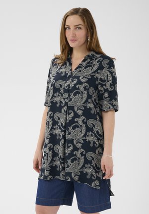 Žena nosi tmavomodrú dlhú košeľu so vzorom paisley a rukávmi do lakťov a rifľové kraťasy po kolená, stojaca pred jednoliatym pozadím.