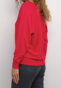 Frau mit lockigem Haar, die einen leuchtend roten Pullover mit langen Ärmeln und dunklen Jeans mit kleinen weißen Punkten trägt, und vor einem einfarbigen Hintergrund steht.