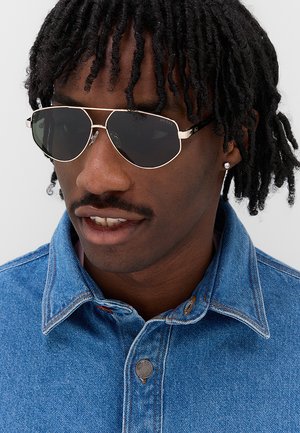 Uomo con dreadlocks corti che indossa occhiali da sole scuri con montatura dorata, un orecchino con diamante e una camicia di jeans blu sbottonata.