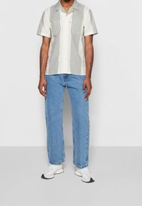 Homme portant une chemise à manches courtes à rayures verticales claires et grises, un jean bleu et des baskets blanches, debout devant un fond uni.