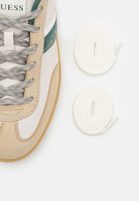 Sneaker with beige suede upper, mesh accents, and a green logo stripe. Includes two white laces with textured detail. Rubber sole.