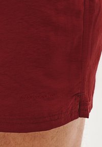 Czerwone szorty wykonane z lekkiego materiału, charakteryzujące się delikatną teksturą i haftowanym logo blisko dołu.