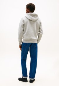 Sudadera con capucha gris de corte holgado, material de algodón y bolsillo tipo canguro, combinada con jeans azules y zapatos negros.