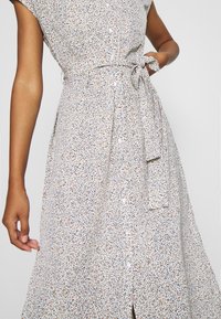 Nainen, jolla on päällään valkoinen, hihaton, napitettava mekko, jossa on pieni kukkakuosi ja sidottu vyö.