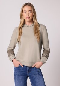 Femme aux longs cheveux blonds portant un sweat à capuche beige avec des poignets à motifs et un jean bleu, debout les mains dans les poches devant un fond uni.