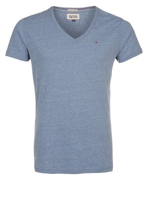 Mand stående med krydsede ben iført en lyseblå V-hals T-shirt, sorte jeans og mørkebrune lædersko mod en ensfarvet baggrund.