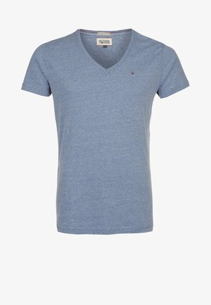 Mann steht mit übereinandergeschlagenen Beinen, trägt ein hellblaues V-Ausschnitt-T-Shirt, schwarze Jeans und dunkelbraune Lederschuhe vor einem einfarbigen Hintergrund.