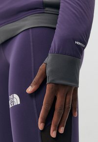 Lilla sportstopp med grå detaljer og en tettsittende passform, med en VENTRIX-logo. Hånden hviler på teksturerte leggings med hvit merkevarelogo.