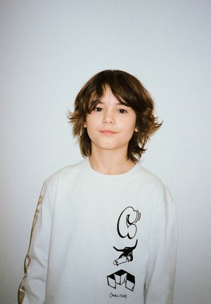 Chłopiec o średniej długości brązowych włosach, noszący białą bluzę z czarnymi wzorami graficznymi i napisem "COALITION" z przodu.