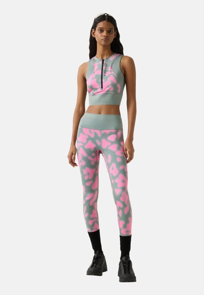 Femme debout portant un crop top sans manches gris et rose à motifs avec une fermeture éclair, un legging assorti, des chaussettes noires et des chaussures épaisses noires.