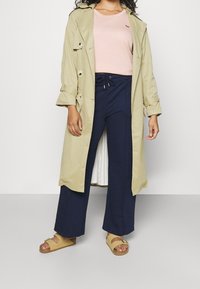 Trench coat beige sopra una maglietta rosa, abbinato a pantaloni ampi blu scuro e sandali beige con fibbia. Tessuto morbido, vestibilità casual.