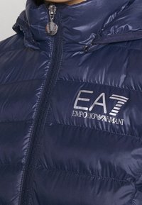 Chaqueta de plumas azul marino con cuello alto, cierre de cremallera y logo EA7 Emporio Armani. Textura suave y brillante con paneles acolchados.