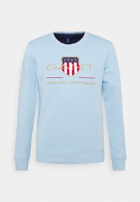 Sudadera azul claro con cuello redondo, puños acanalados y un logo con un diseño de escudo con rayas rojas y blancas y bordado dorado.