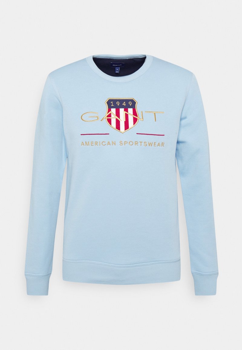 Sudadera azul claro con cuello redondo, puños acanalados y un logo con un diseño de escudo con rayas rojas y blancas y bordado dorado.