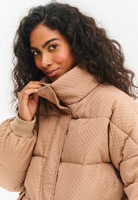 Prešívaná bunda svetlohnedej farby s textúrovaným povrchom, vysokým golierom a rebrovanými manžetami. Má prešívané časti a voľný strih.