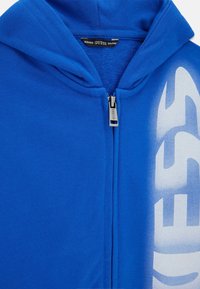 La sudadera con cremallera en azul royal presenta una textura suave, capucha y un logotipo blanco impreso en la parte delantera. Cremallera metálica con detalle de marca.