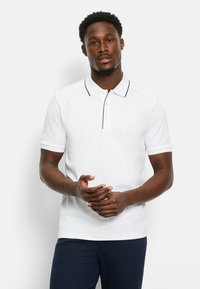 Weißes Poloshirt mit einer Knopfleiste mit zwei Knöpfen, schwarzer Paspelierung entlang des Kragens, kurzen Ärmeln und glatter Textur. Getragen mit dunkelblauen Hosen.