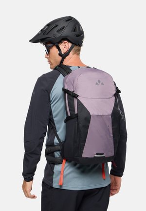 Man die een zwarte fietshelm, zonnebril en een grijs-blauwe jas met een paarse en zwarte rugzak draagt, tegen een effen achtergrond.