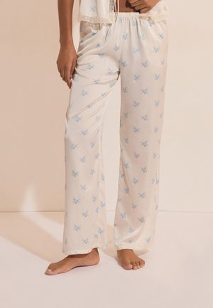 Person trägt lockere cremefarbene Pyjamahosen mit kleinem blauem Blumenmuster, steht barfuß vor einem beigen Hintergrund.