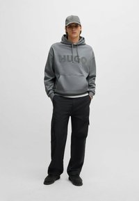 Grijze hoodie met "HUGO" logo, trekkoord capuchon en kangoeroezak. Gecombineerd met zwarte cargobroek en een grijze pet. Zwart schoeisel.