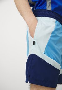 Herre sportsshortser i et lagdelt design, med lyseblå og marineblå paneler, sidelommer og et stoffmerke på sømmen.