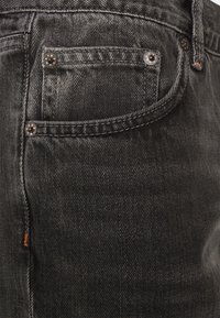 Vaqueros de mezclilla negra con una vista en primer plano que muestra un bolsillo delantero, remaches metálicos, costuras en contraste y una textura ligeramente desgastada.