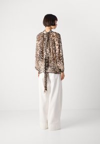 Femme aux cheveux courts portant une blouse en dentelle imprimée léopard nouée au cou et un large pantalon blanc, se tenant devant un fond uni.