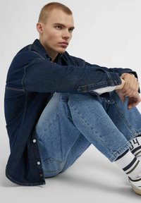 Camicia di denim blu scuro con cuciture a contrasto, indossata sopra jeans di denim blu chiaro. Calze bianche a righe completano l'outfit.