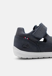 Tmavě modrá kožená bota s prodyšným designem, suchým zipem, bílou podešví a červenými, bílými a modrými akcenty na patě.