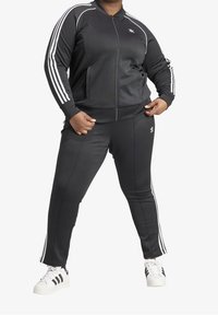 Černá tepláková souprava s bílými pruhy po stranách, obsahující bundu na zip a přiléhavé kalhoty, vyrobená z hladké látky. Outfit doplňují tenisky.