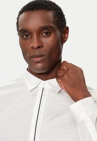 Camicia bianca con collo a punta, caratterizzata da una striscia verticale nera e un piccolo logo ricamato sul lato sinistro.